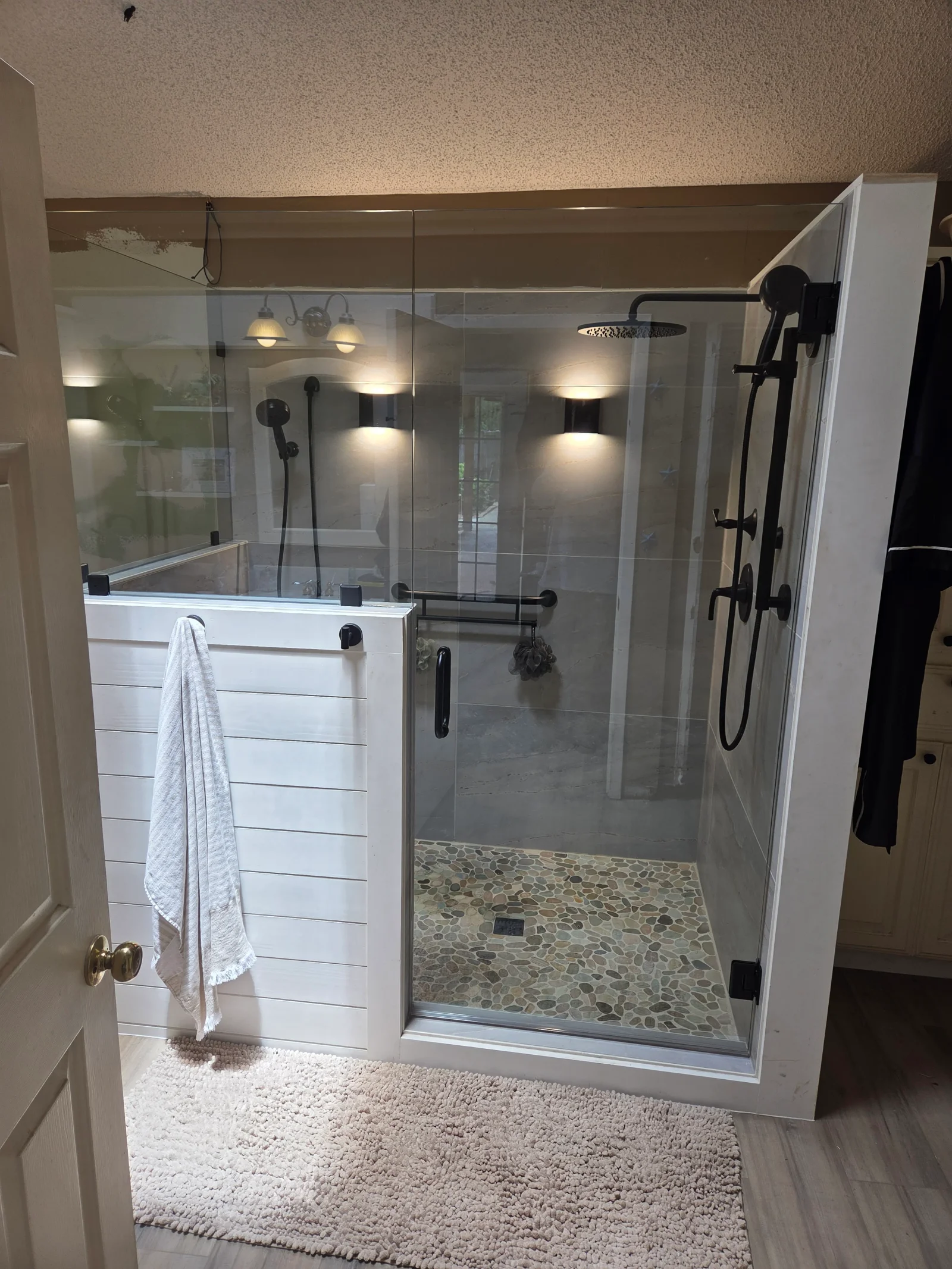 Frameless glass shower enclosure with pebble floor, white shiplap vanity, black fixtures, and recessed lighting