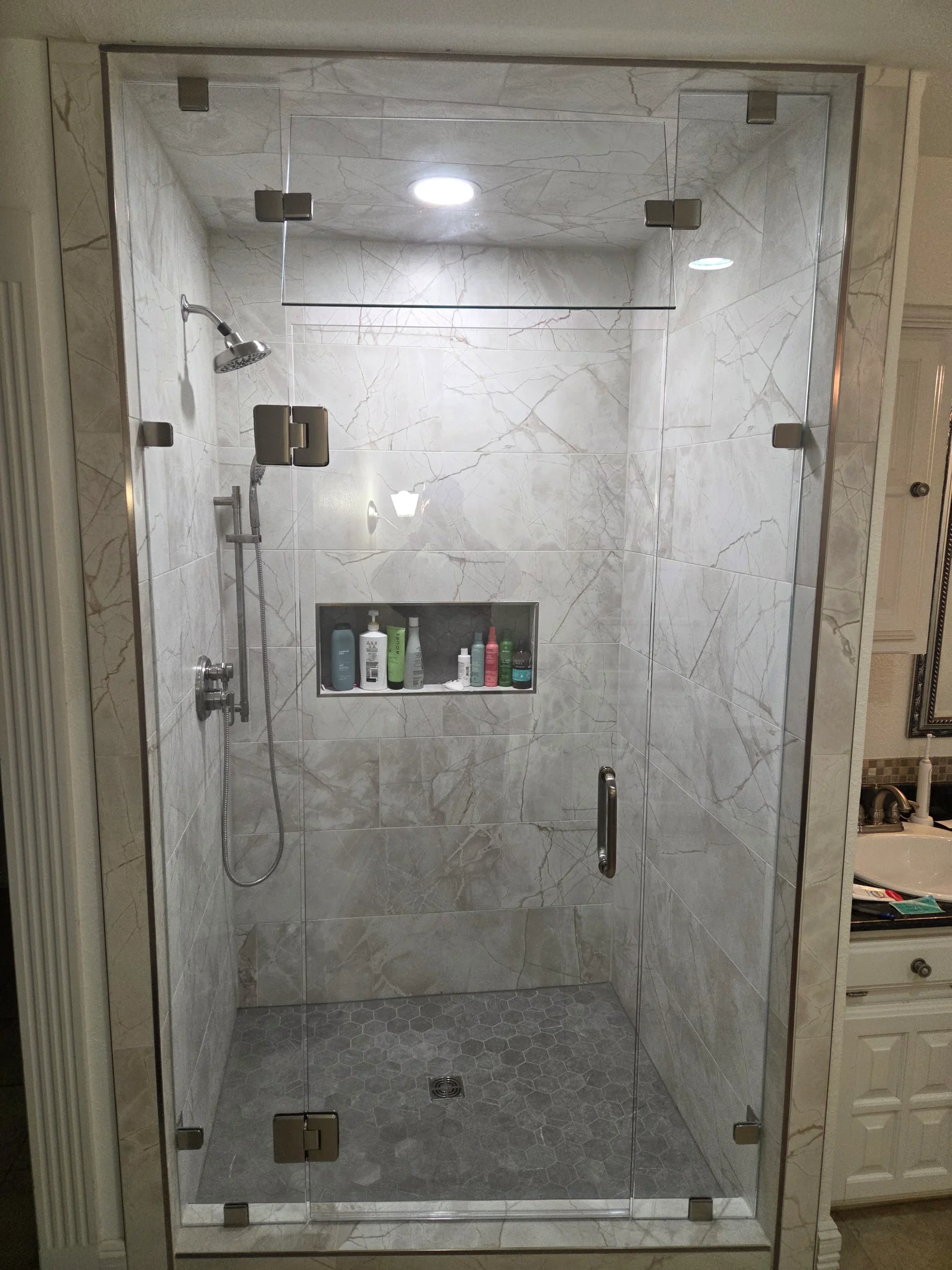 Marble tile shower with frameless glass doors, rainfall showerhead, and recessed storage niche
