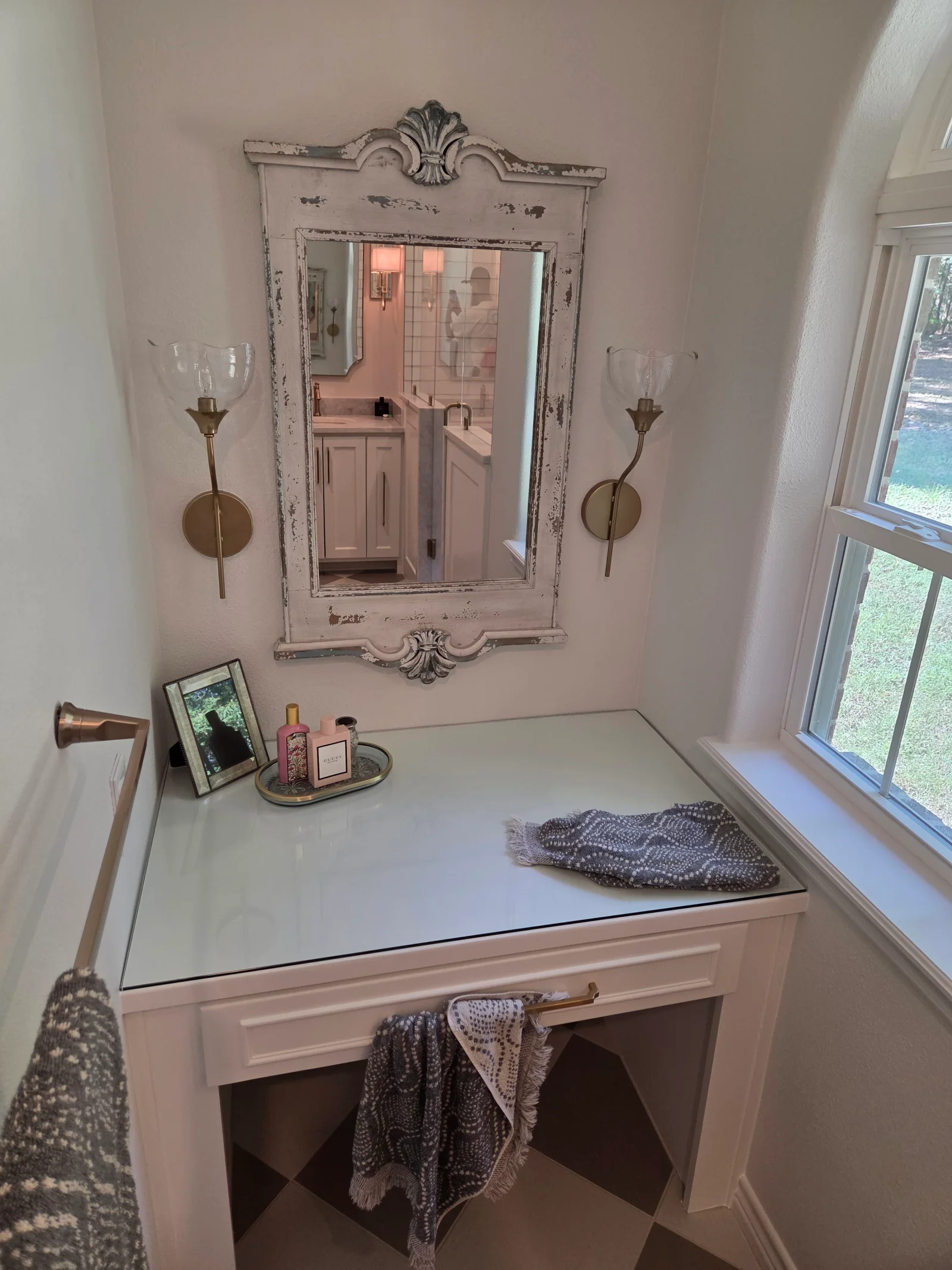 White quartz vanity with distressed wood-framed mirror, brass sconces, and pink cabinetry