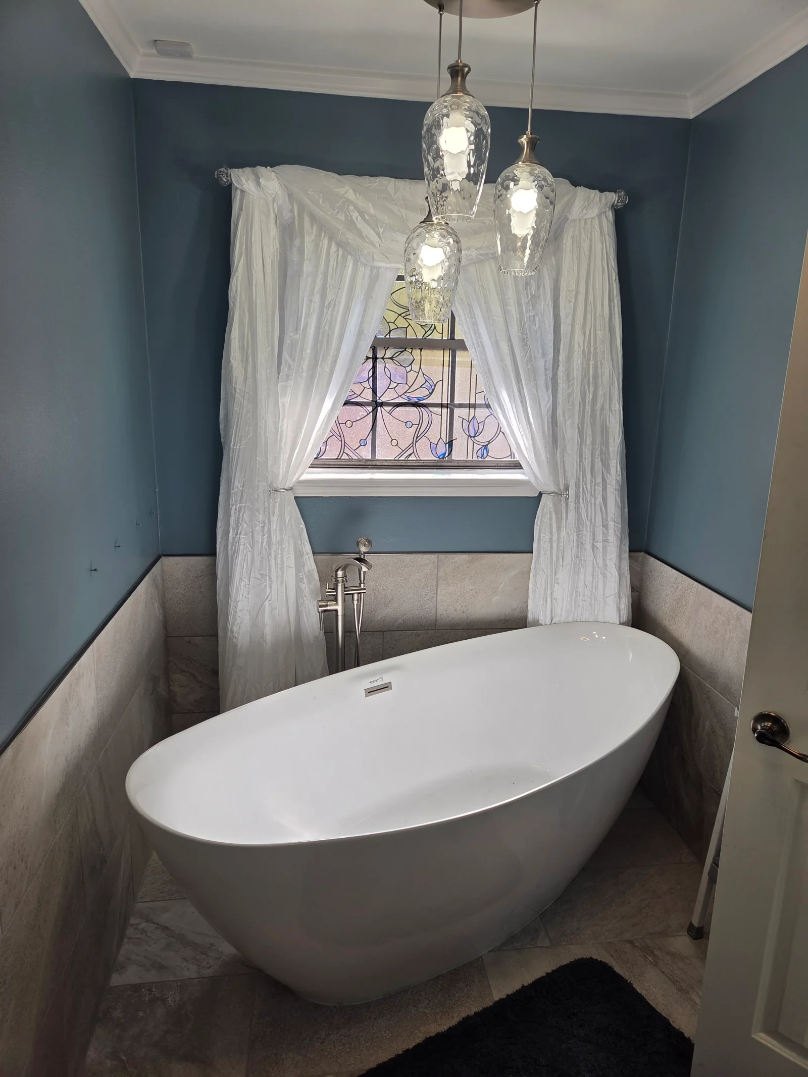 Freestanding white tub, gray tile wainscoting, teal walls, pendant lighting, stained glass window